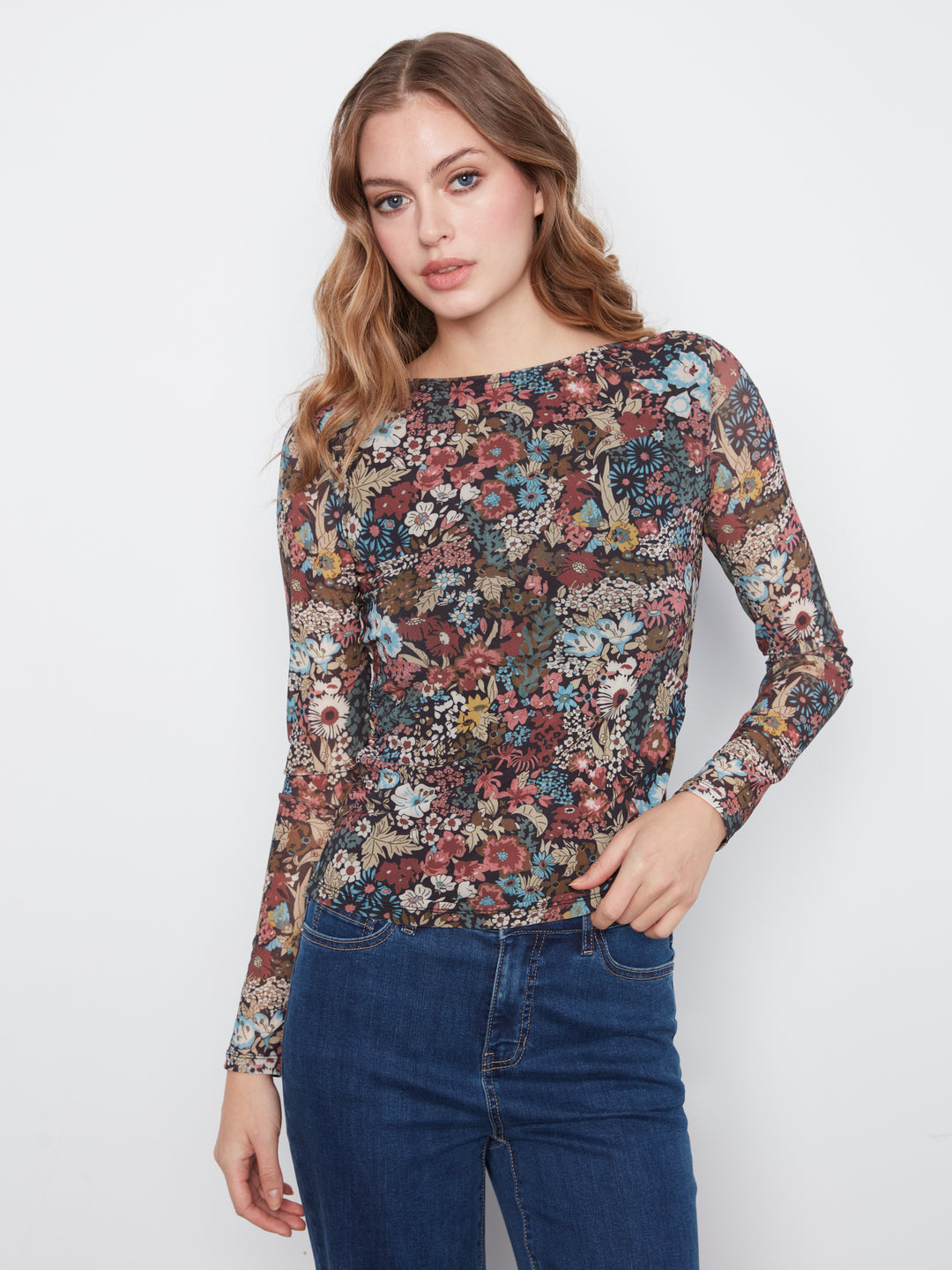 A woman modeling a long-sleeve boat-neck top with an intricate multicolor floral and bird pattern, paired with dark blue jeans, against a plain light gray background.