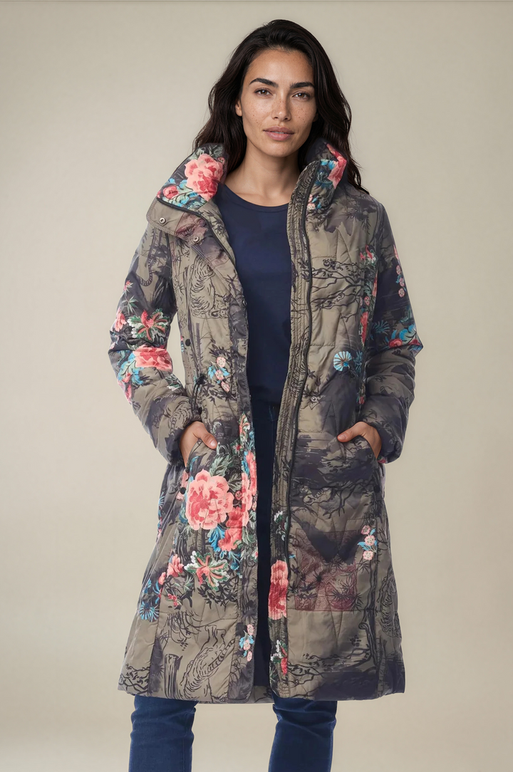 A woman modeling a long quilted puffer coat with a floral and scenic print. The coat features a mix of pink, blue, green, and brown tones, with a large floral pattern on the hood and pockets.