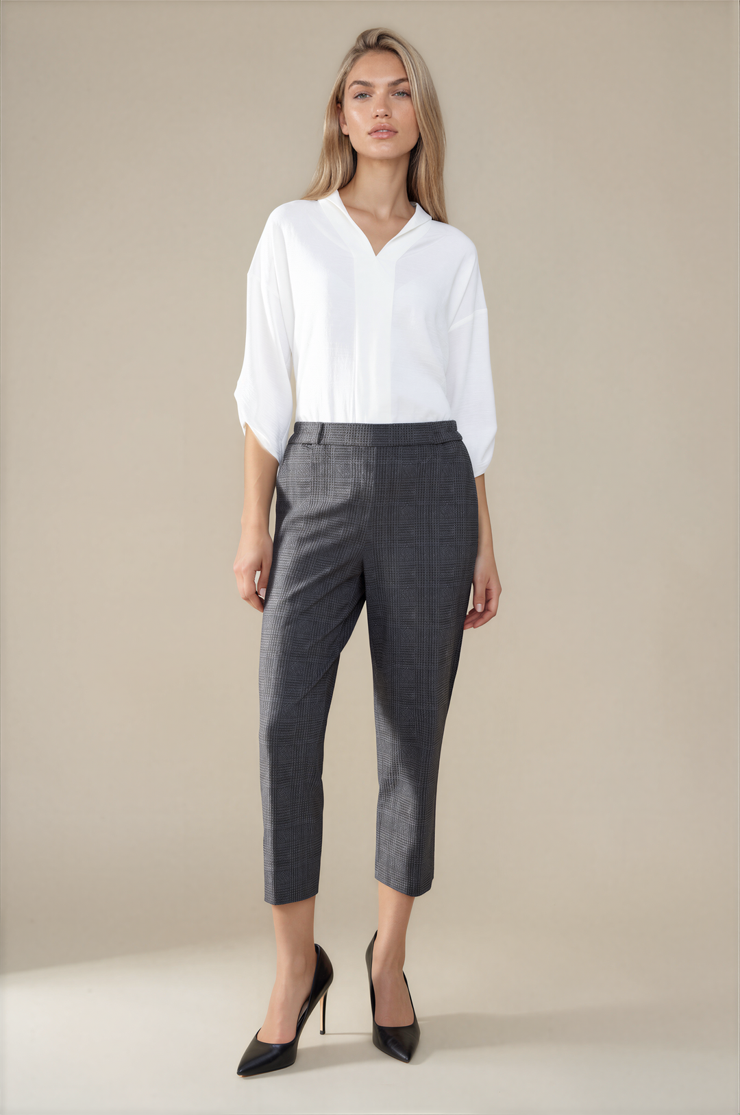 A woman stands against a plain beige studio background wearing a white blouse, dark gray cropped trousers, and black pointed heels.