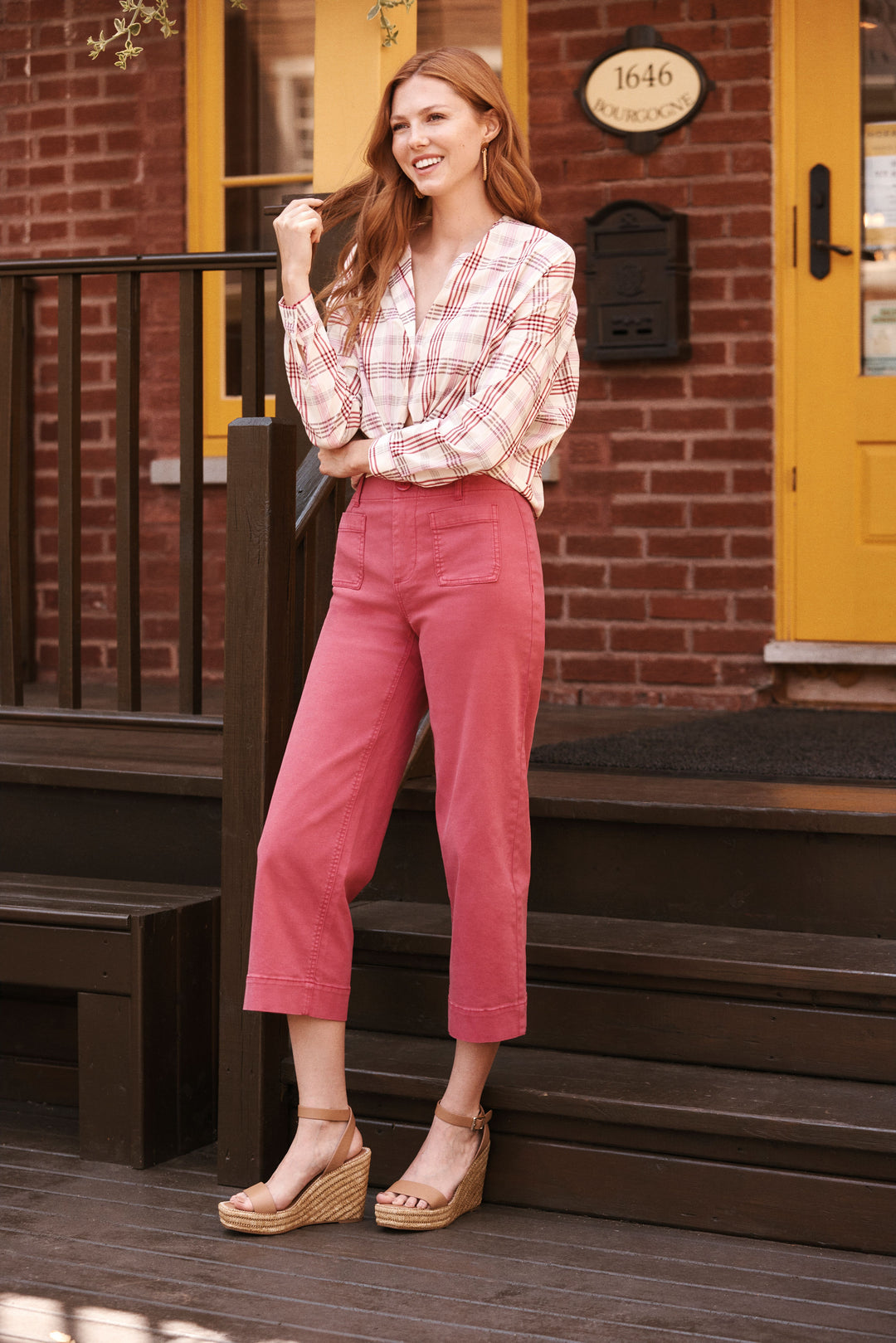 Woman in a plaid shirt and pink pants standing on a wooden porch with a yellow door in the background.