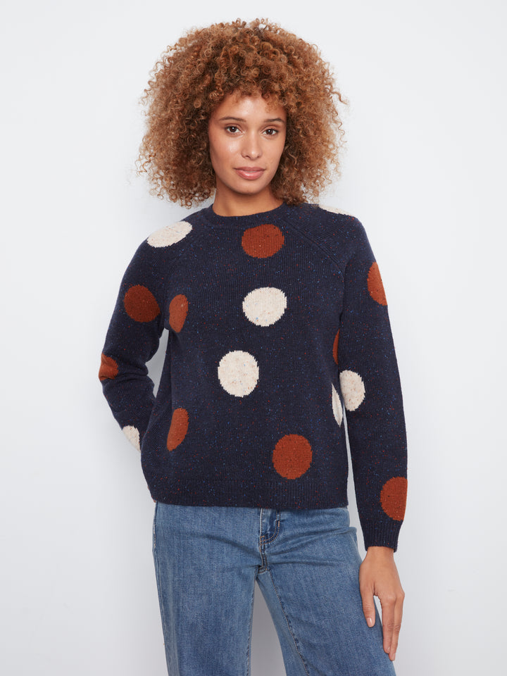 A woman with curly brown hair wearing a navy blue knit sweater with large textured pom-pom polka dots in rust orange and cream, paired with blue jeans, against a plain white studio background.