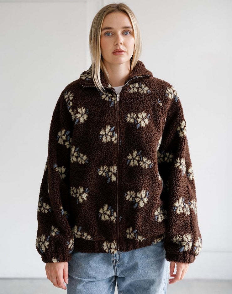 A woman wearing a brown sherpa fleece zip-up jacket with a cream and blue floral pattern, paired with light blue jeans, standing against a plain white wall.