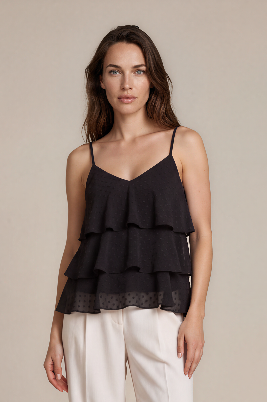 A woman modeling a black ruffled camisole top with thin straps and white wide-leg trousers, standing against a plain beige studio background.