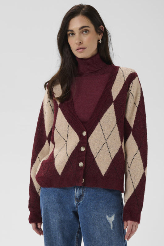 A woman modeling a burgundy and beige argyle cardigan over a matching burgundy turtleneck sweater, paired with blue jeans, against a light gray studio background.