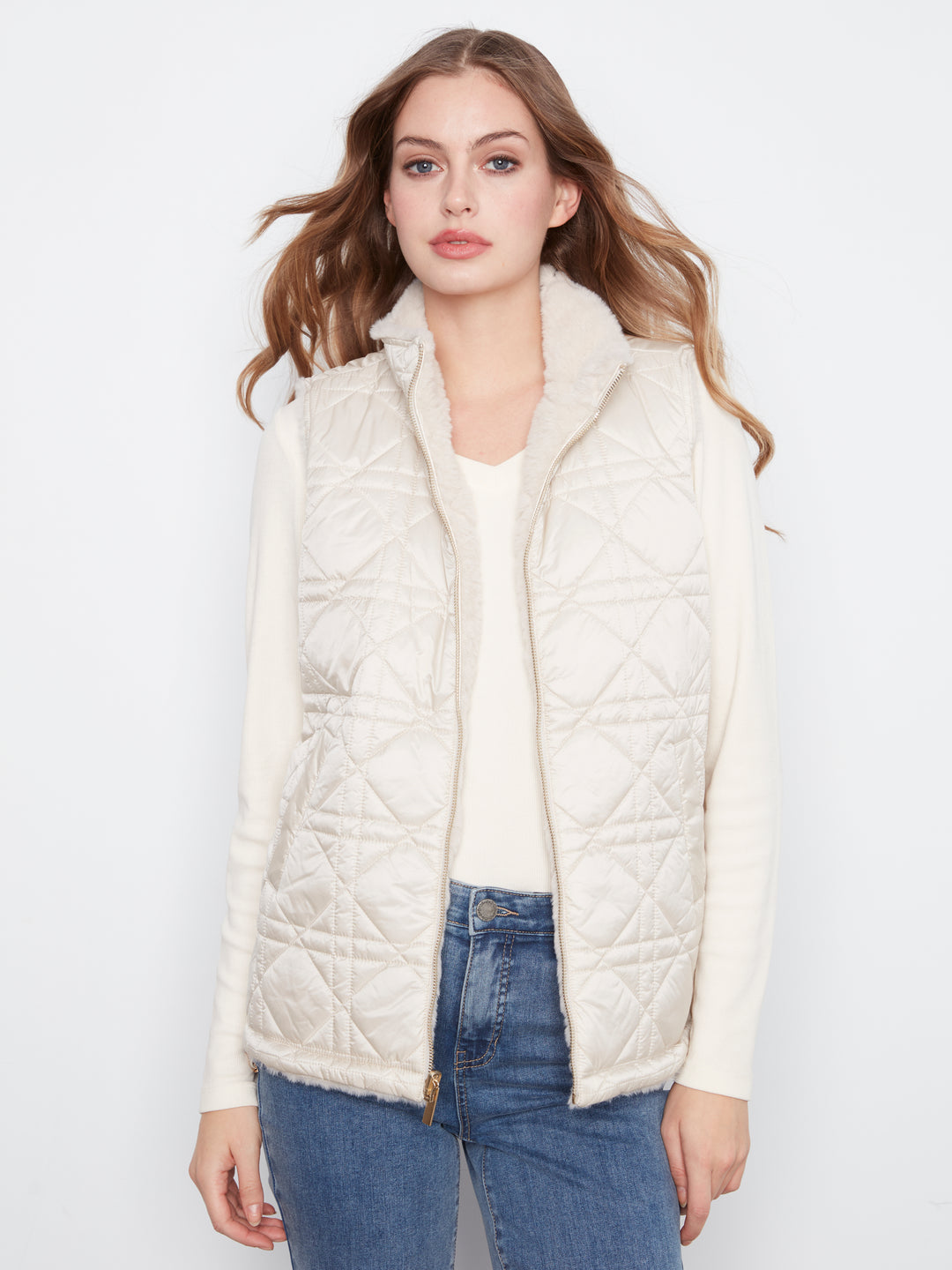 A woman modeling a cream-colored quilted puffer vest with a fuzzy collar, worn over a matching cream long-sleeve top and blue jeans, against a plain white studio background.