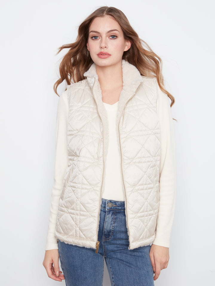 A woman modeling a cream-colored quilted puffer vest with a fuzzy collar, worn over a matching cream long-sleeve top and blue jeans, against a plain white studio background.