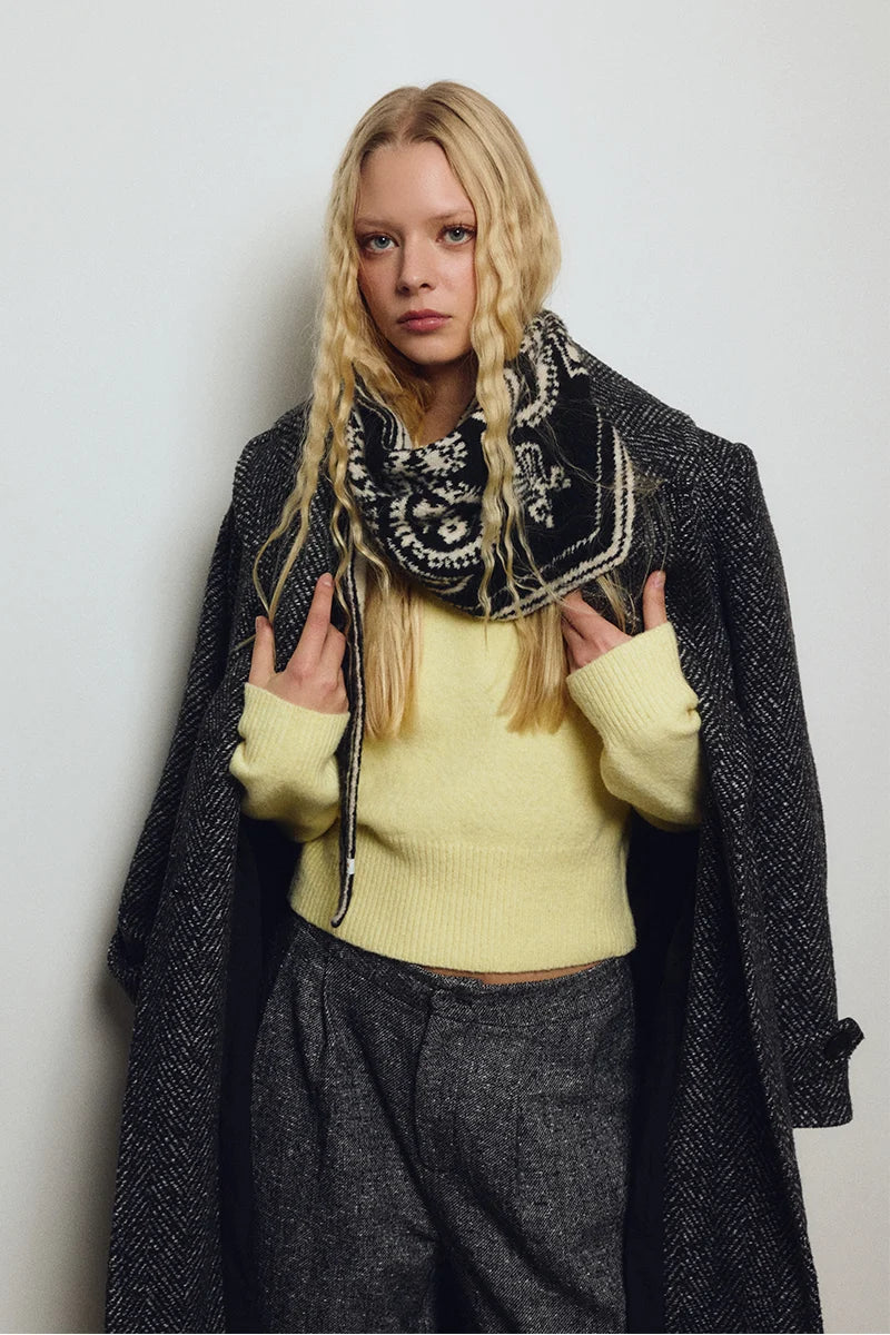 A blonde model wearing a pale yellow knit sweater, a black and white patterned bandana scarf, and a dark gray textured overcoat, posing against a light gray background.