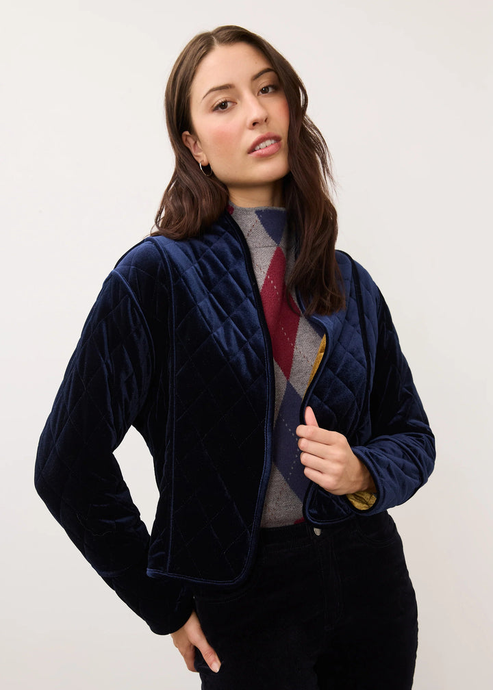 A woman modeling a navy blue velvet cropped quilted jacket over a gray and red argyle sweater, paired with black pants, against a plain off-white studio background.