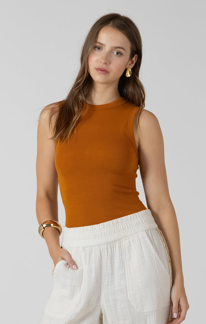 Woman wearing a mustard yellow sleeveless top and white pants on a plain background
