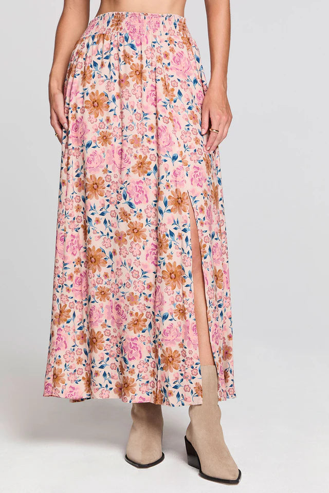 A model wears a high-waisted, pleated maxi skirt with a pink, orange, and blue floral pattern and a front slit, paired with beige ankle boots against a light gray studio background.