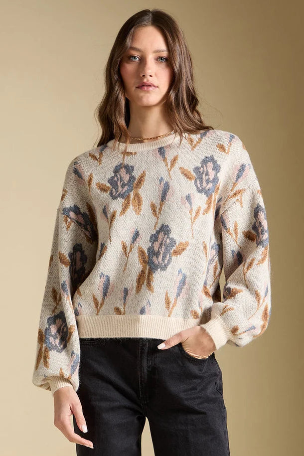 A woman modeling a cream-colored knit sweater with a blue and brown floral pattern, paired with black jeans, against a solid beige background.