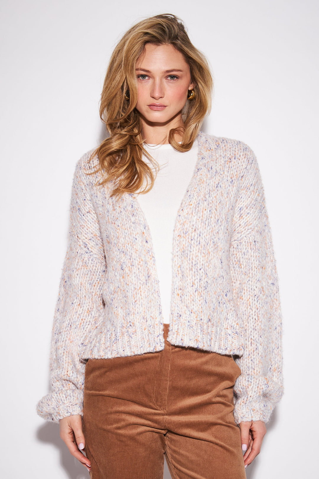 A woman modeling a cream-colored, textured open-front cardigan with subtle multicolor flecks, worn over a white top and brown corduroy pants, against a plain white studio background.