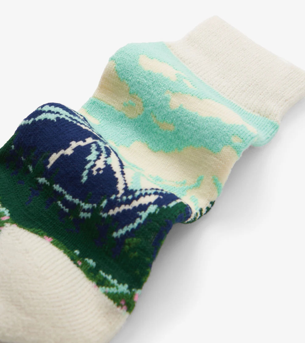 A close-up of a single knit sock with a scenic landscape pattern featuring a dark blue sky, green hills, and cream clouds, set against a plain white background.