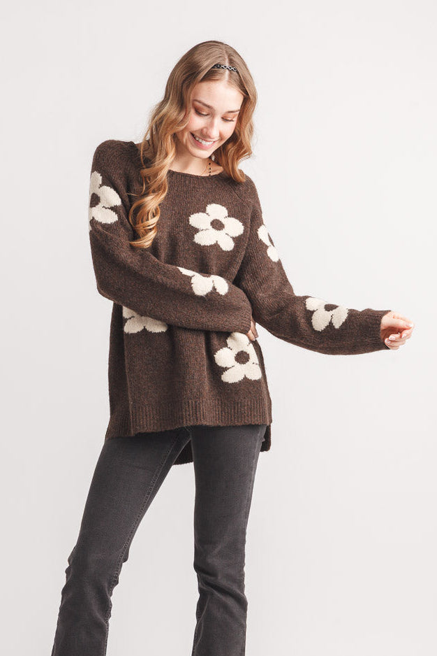 A woman modeling a brown knit sweater with large white flower motifs on the sleeves and body, paired with dark gray pants and black shoes, against a light gray studio background.