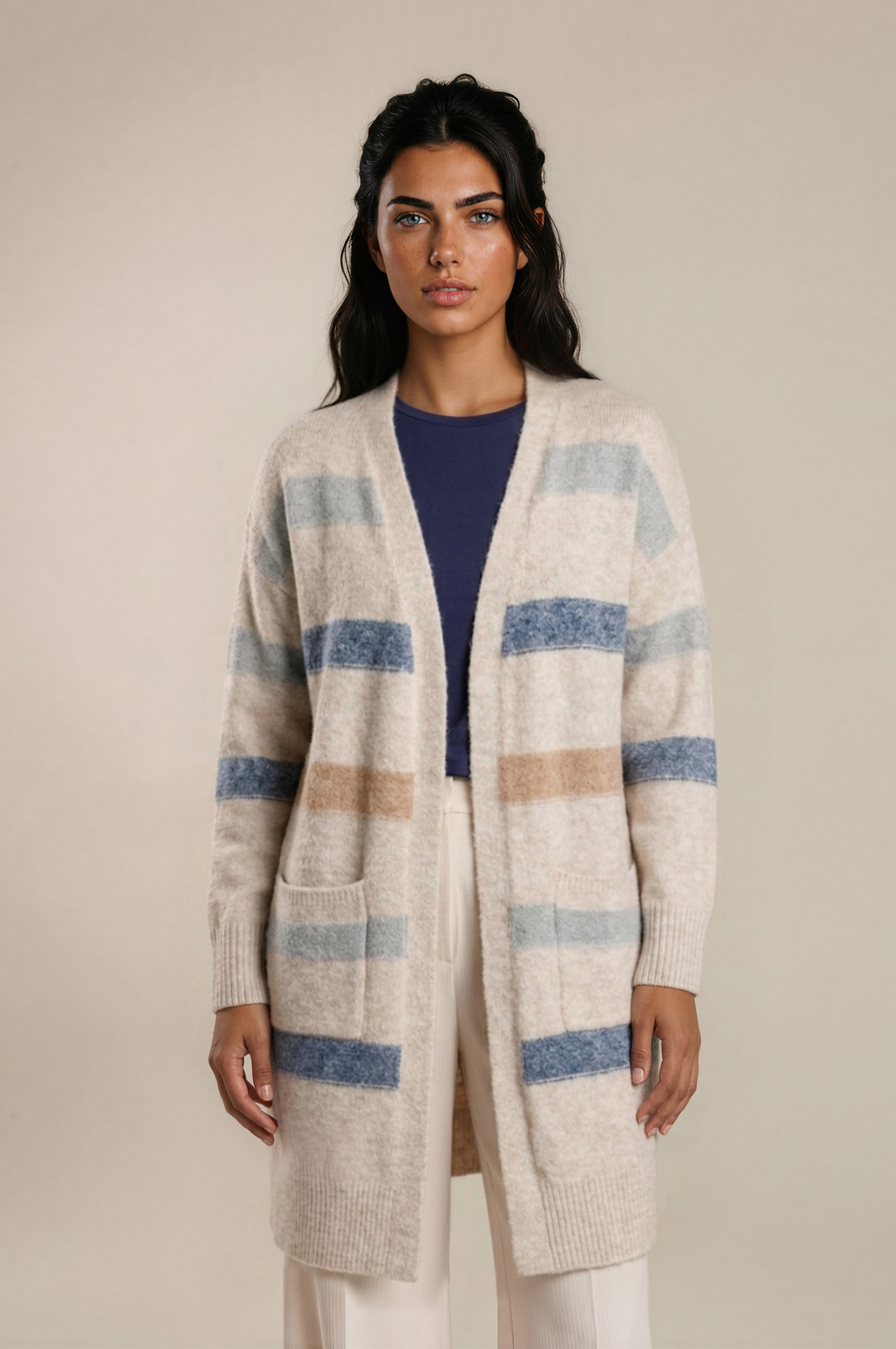 A woman modeling a long open-front knit cardigan with horizontal stripes in blue, beige, and light gray on an off-white base, worn over a navy top and cream pants against a plain beige studio background.