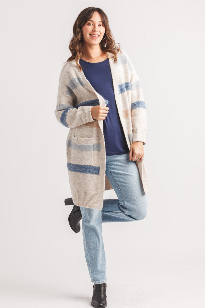 A woman modeling a long open-front cardigan with a beige, blue, and white plaid pattern, worn over a navy top and light blue jeans, against a plain light gray studio background.