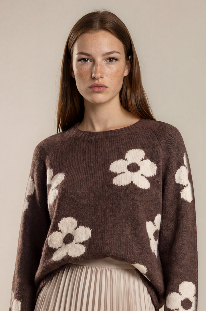 A woman modeling a brown knit sweater with large white floral patterns, paired with a pleated beige midi skirt, against a plain beige studio background.