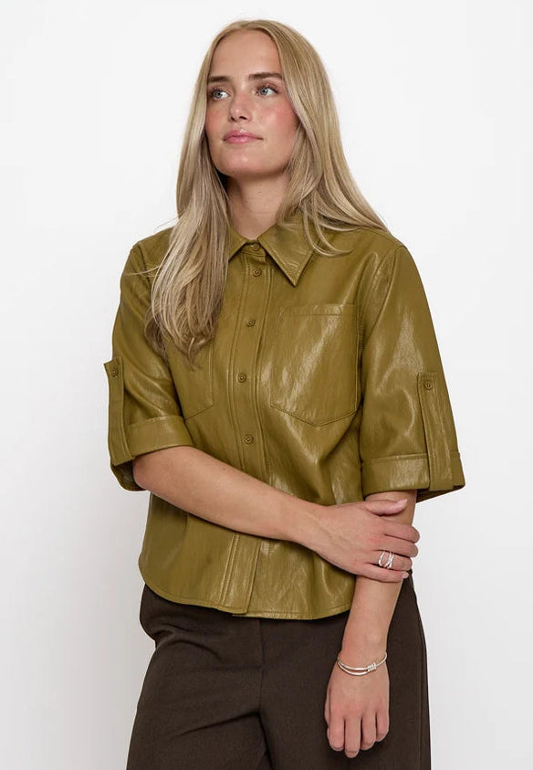 A blonde woman wearing a short-sleeved olive green faux leather button-up shirt and dark brown trousers, standing against a plain white studio background.