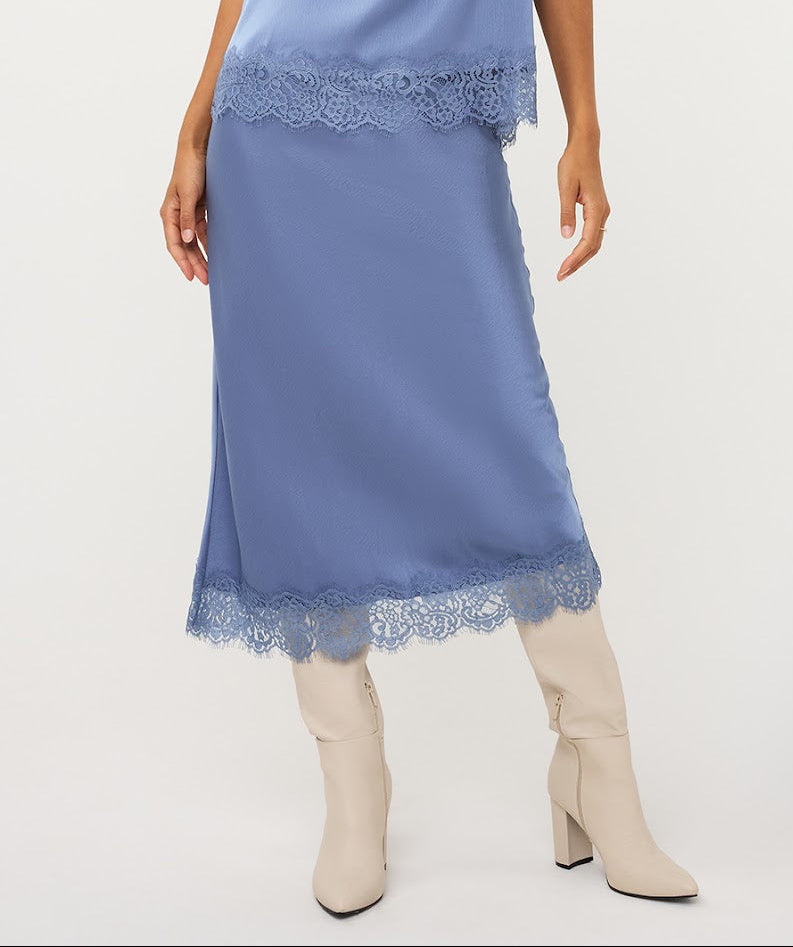 A model is wearing a dusty blue satin midi skirt with matching blue lace trim at the waist and hem, paired with beige heeled ankle boots against a plain white studio background.