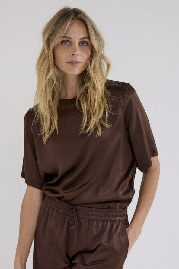 A woman modeling a matching brown lounge set consisting of a short-sleeve top and drawstring pants, photographed against a plain light gray studio background.