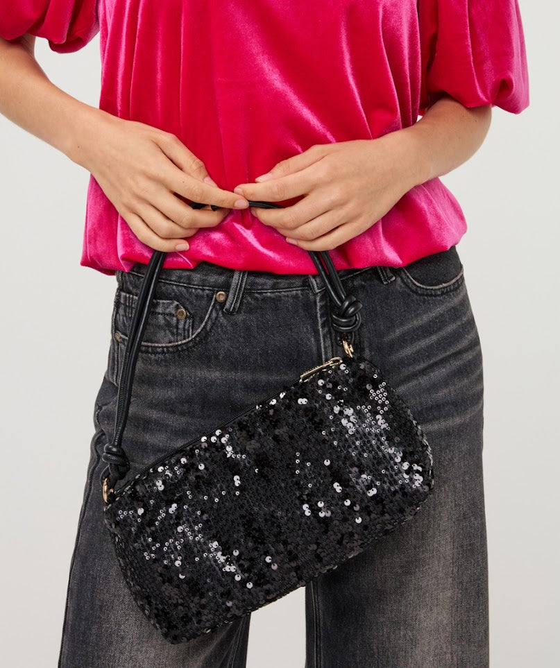 A person wearing a bright pink velvet top and dark gray jeans holds a black sequined clutch bag with a black strap against a light gray studio background.