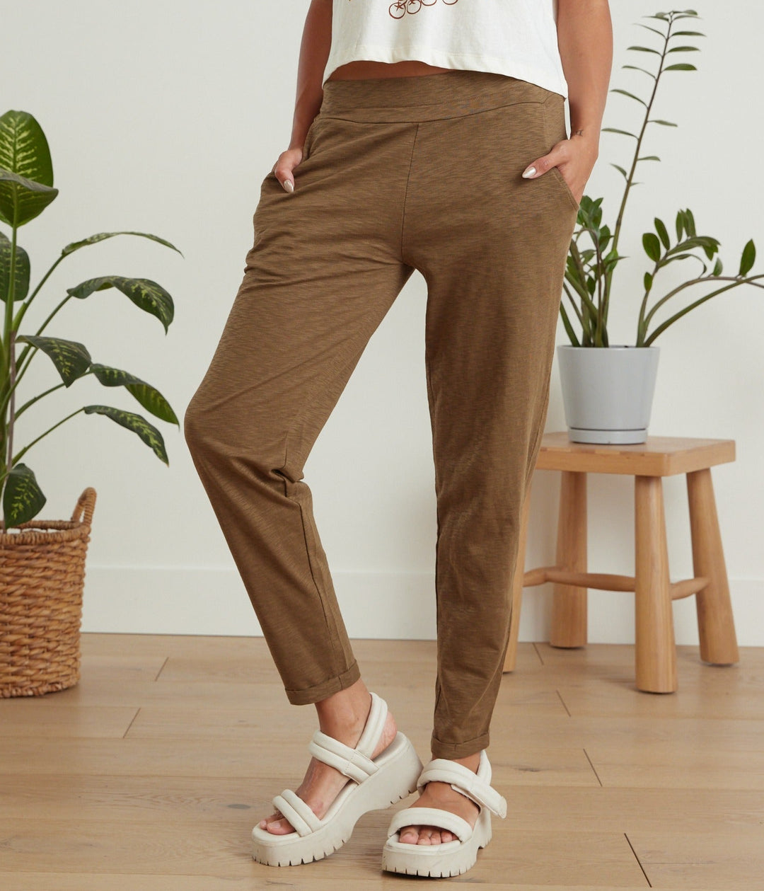 A person wearing brown cropped pants with a white graphic t-shirt and white platform sandals, standing in a room with wooden floors, potted plants, and a small wooden stool.