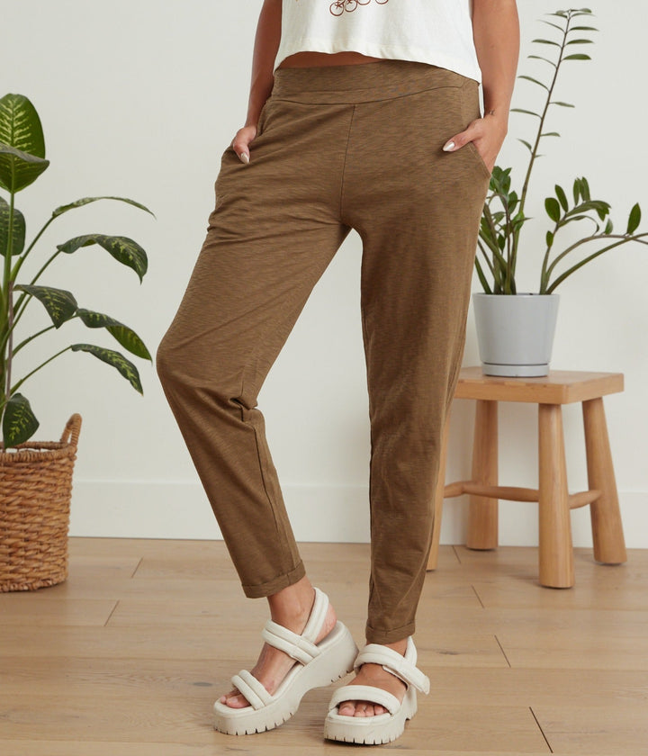 A person wearing brown cropped pants with a white graphic t-shirt and white platform sandals, standing in a room with wooden floors, potted plants, and a small wooden stool.