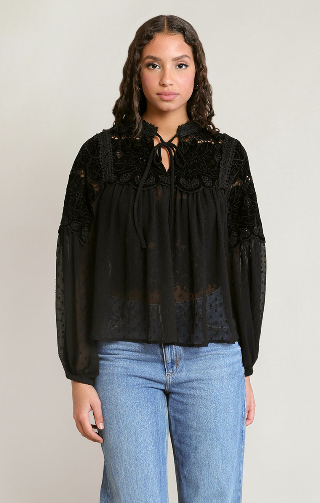 A woman modeling a black sheer lace blouse with long sleeves and a tie-neck detail, paired with light blue jeans, against a plain light gray studio background.