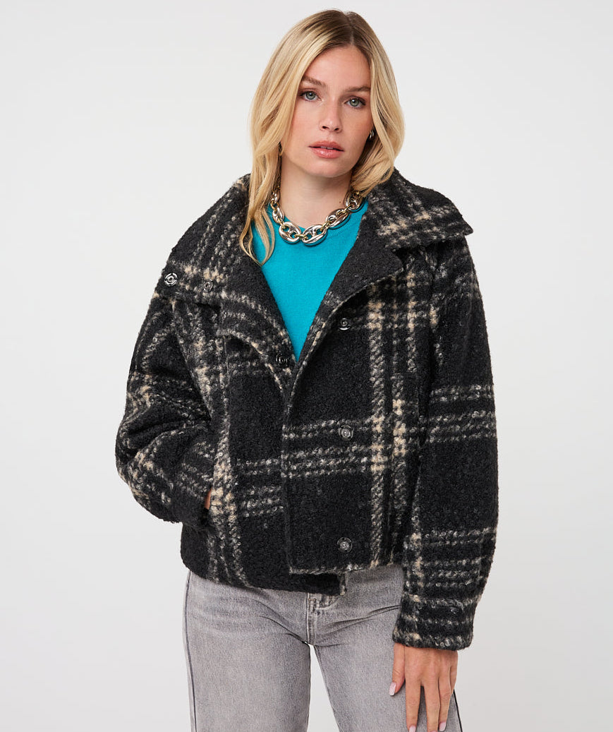 A woman modeling a black and beige plaid sherpa jacket over a bright turquoise top and gray jeans, photographed against a plain light gray studio background.
