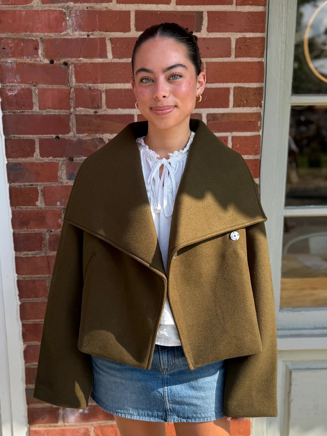 A woman stands outdoors in front of a red brick building, wearing a wide-brimmed olive green wrap coat over a white blouse and denim skirt.