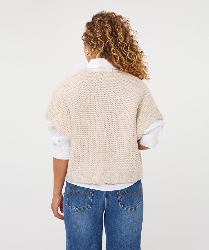 A woman viewed from the back wearing a cream-colored textured knit cardigan over a white collared shirt and blue jeans, against a plain white studio background.