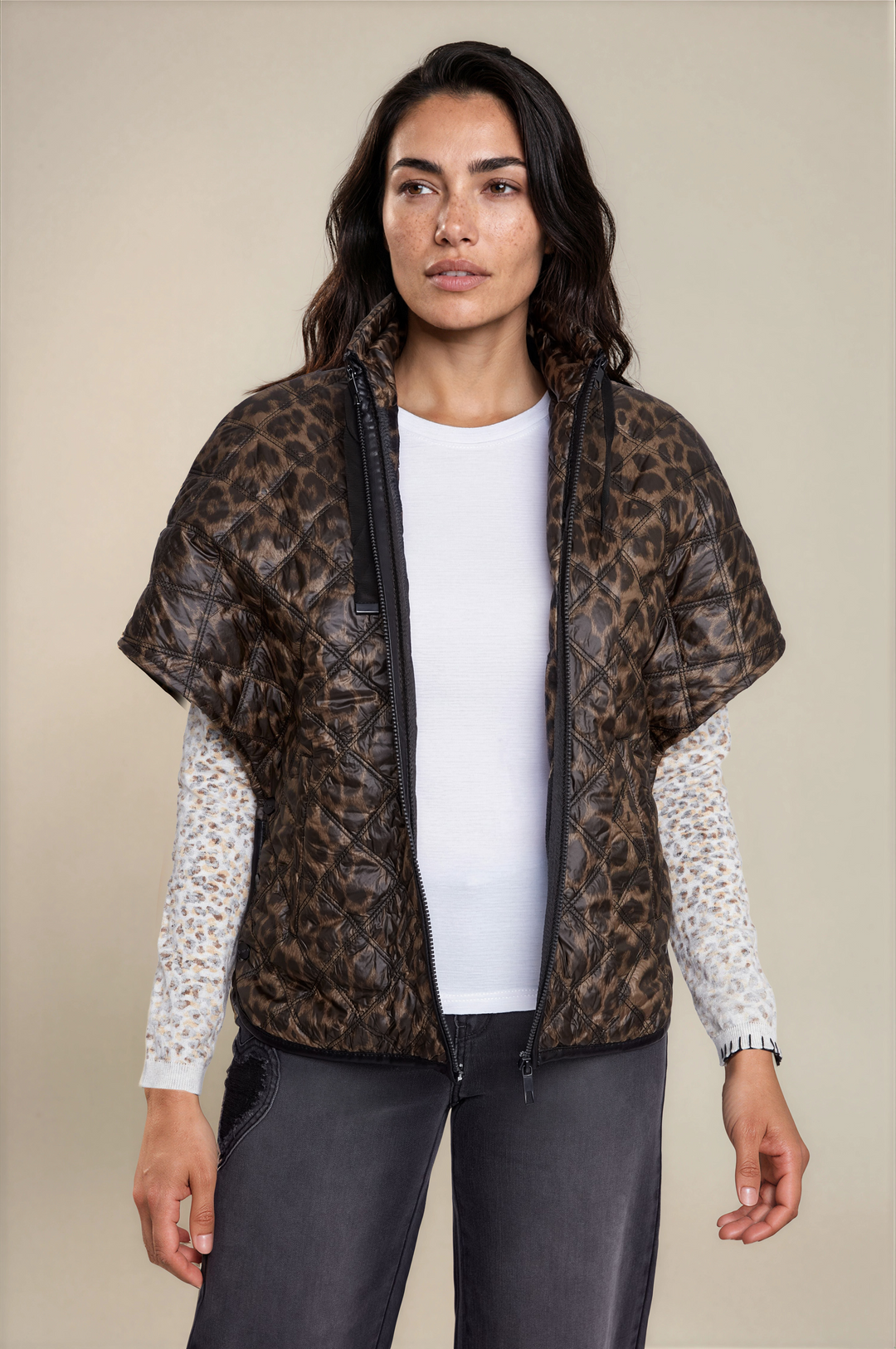 A woman modeling a leopard-print quilted poncho-style jacket over a white long-sleeve top and dark gray pants, photographed against a plain beige studio background.