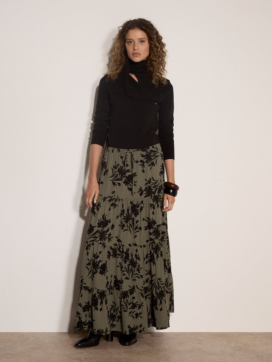 A model stands against a plain off-white wall wearing a black long-sleeve top with a keyhole cutout and a long olive green skirt featuring a black floral pattern.