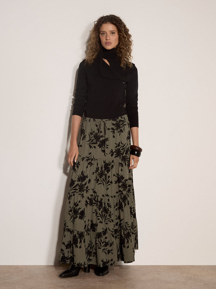 A model stands against a plain off-white wall wearing a black long-sleeve top with a keyhole cutout and a long olive green skirt featuring a black floral pattern.