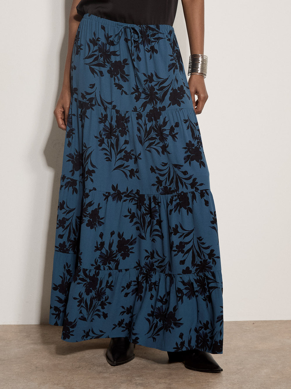A model wearing a long, tiered maxi skirt in a dusty blue fabric with a black floral pattern, paired with black pointed-toe shoes and a silver cuff bracelet.