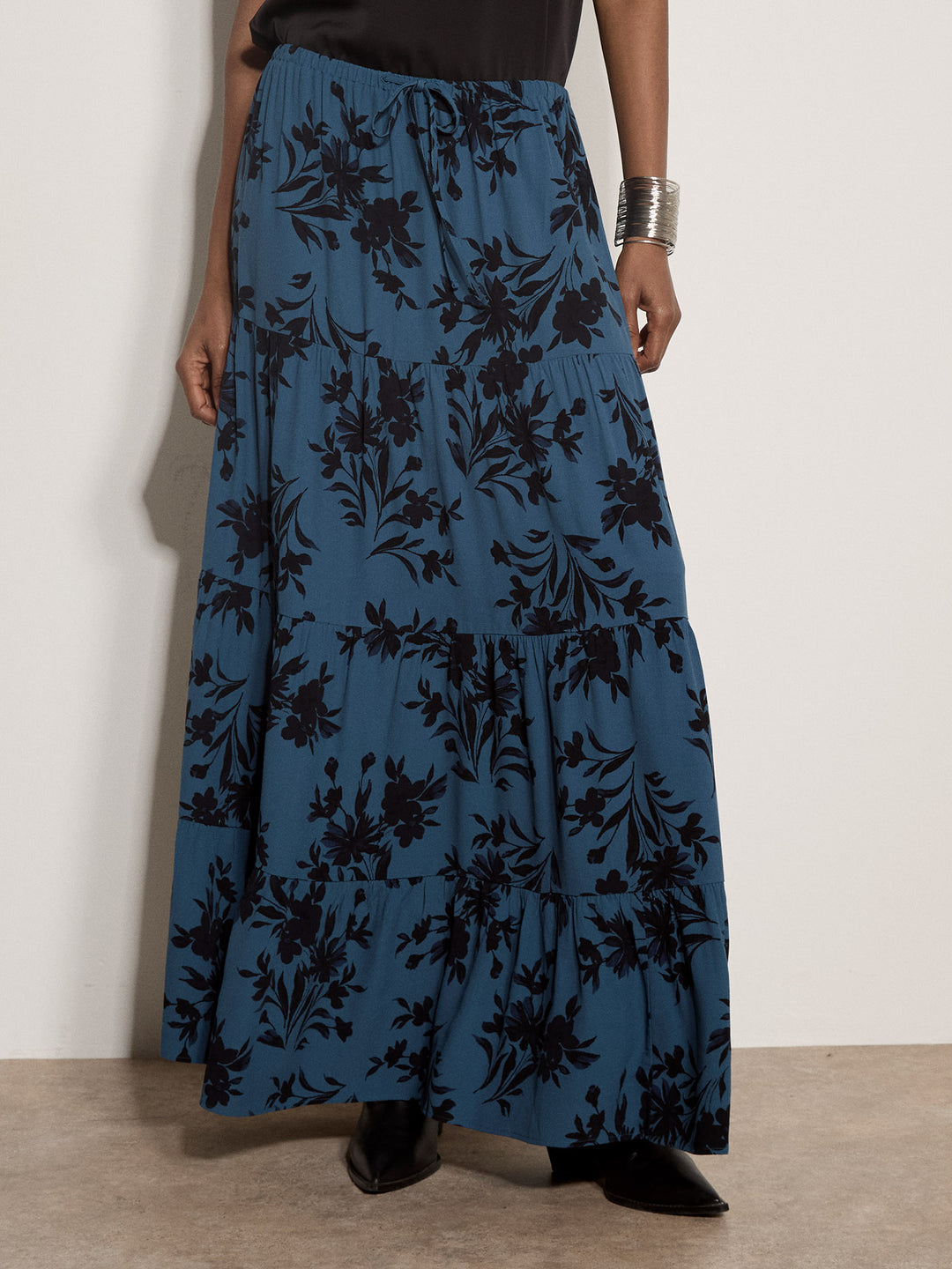 A model wearing a long, tiered maxi skirt in a dusty blue fabric with a black floral pattern, paired with black pointed-toe shoes and a silver cuff bracelet.