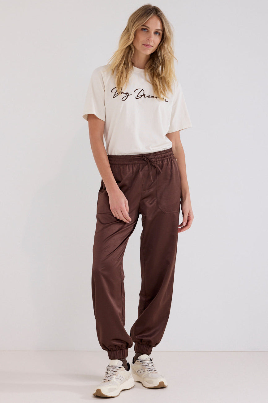 A woman modeling a white graphic t-shirt and dark brown jogger pants with white sneakers, standing against a plain white studio background.
