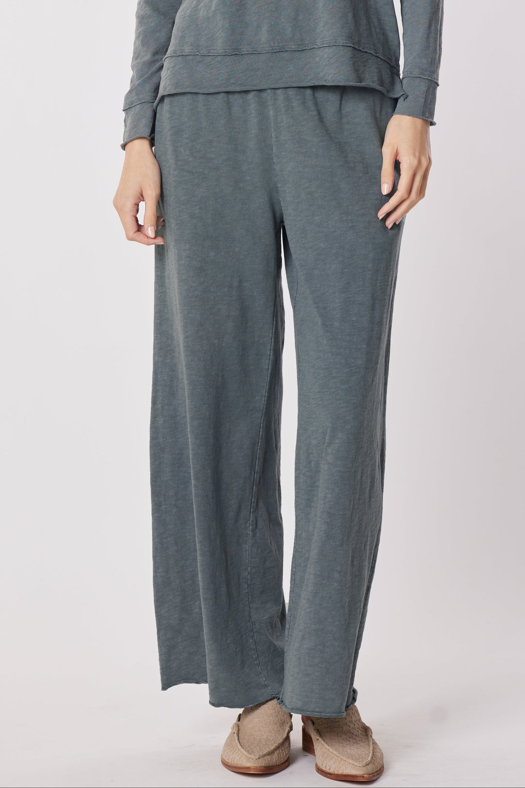 A woman modeling a matching two-piece lounge set consisting of a long-sleeve crewneck sweatshirt and wide-leg pants in a heathered slate gray color, paired with tan slip-on shoes, against a plain light gray studio background.