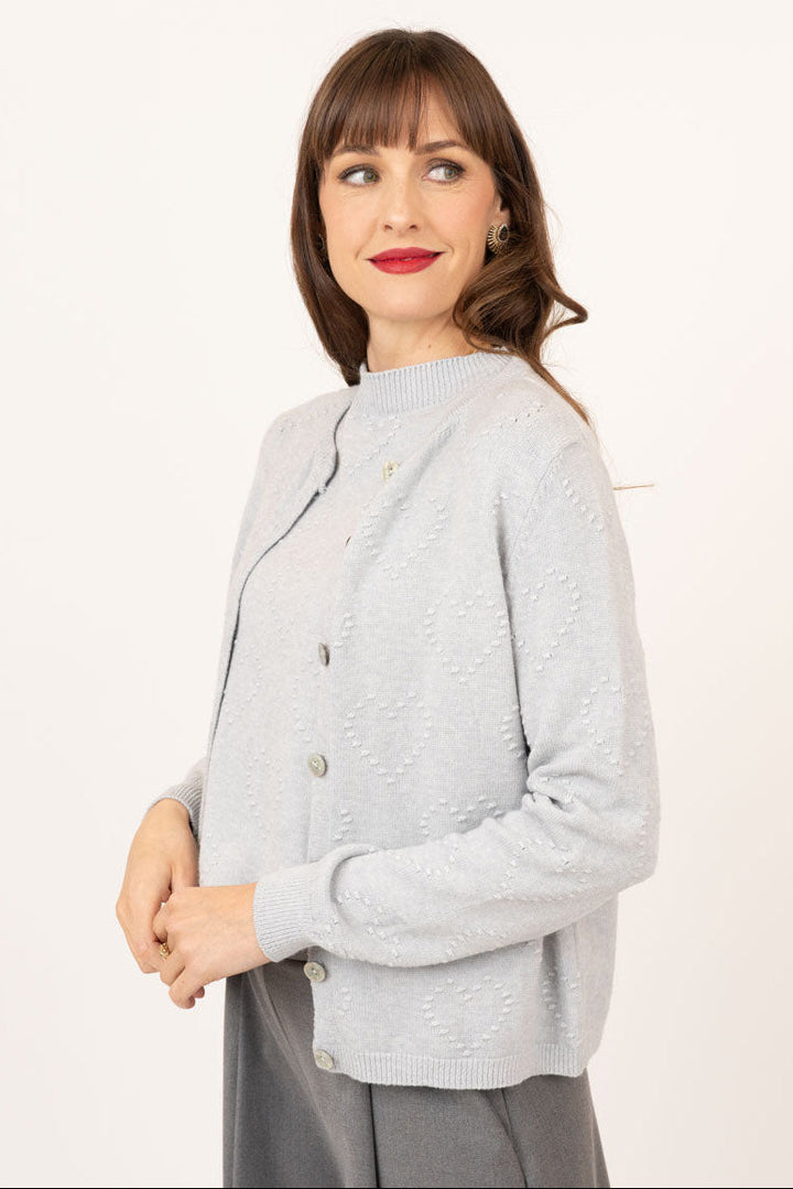 A woman modeling a light gray textured knit cardigan with a mock neck and button closure, paired with dark gray wide-leg trousers, against a plain white studio background.