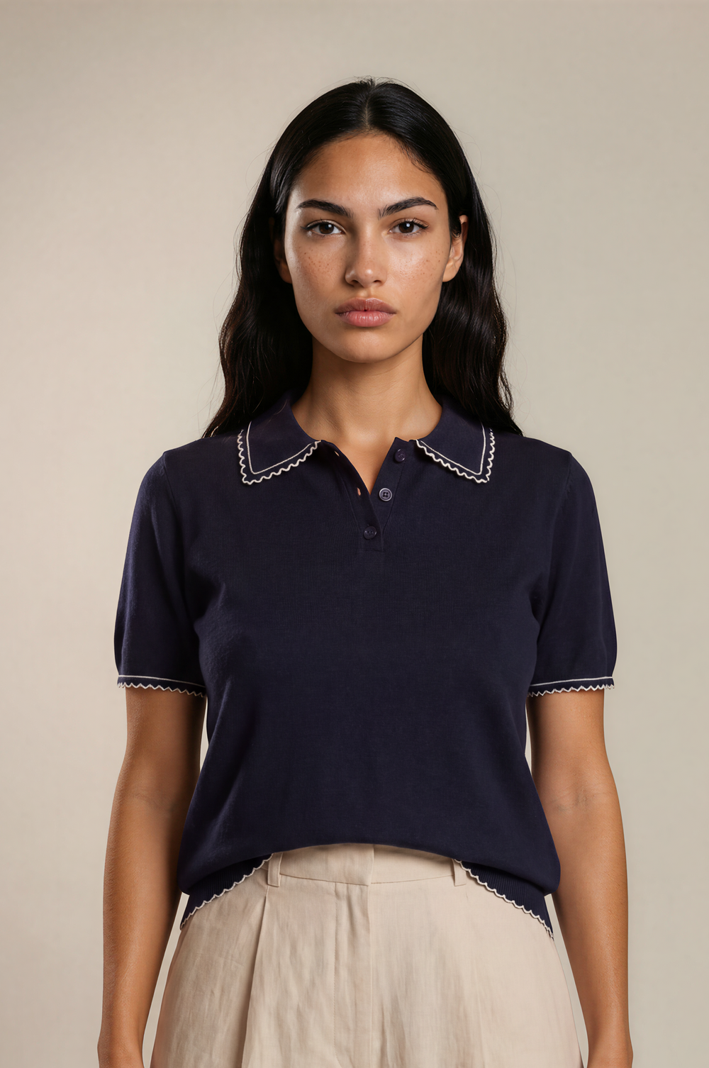 A woman modeling a navy blue short-sleeve polo shirt with white embroidered trim on the collar and sleeve cuffs, paired with light beige linen shorts, against a solid beige studio background.