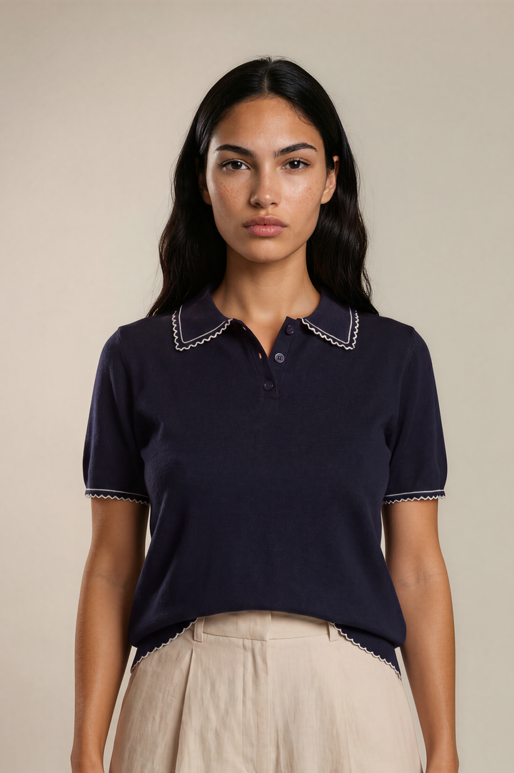 A woman modeling a navy blue short-sleeve polo shirt with white embroidered trim on the collar and sleeve cuffs, paired with light beige linen shorts, against a solid beige studio background.