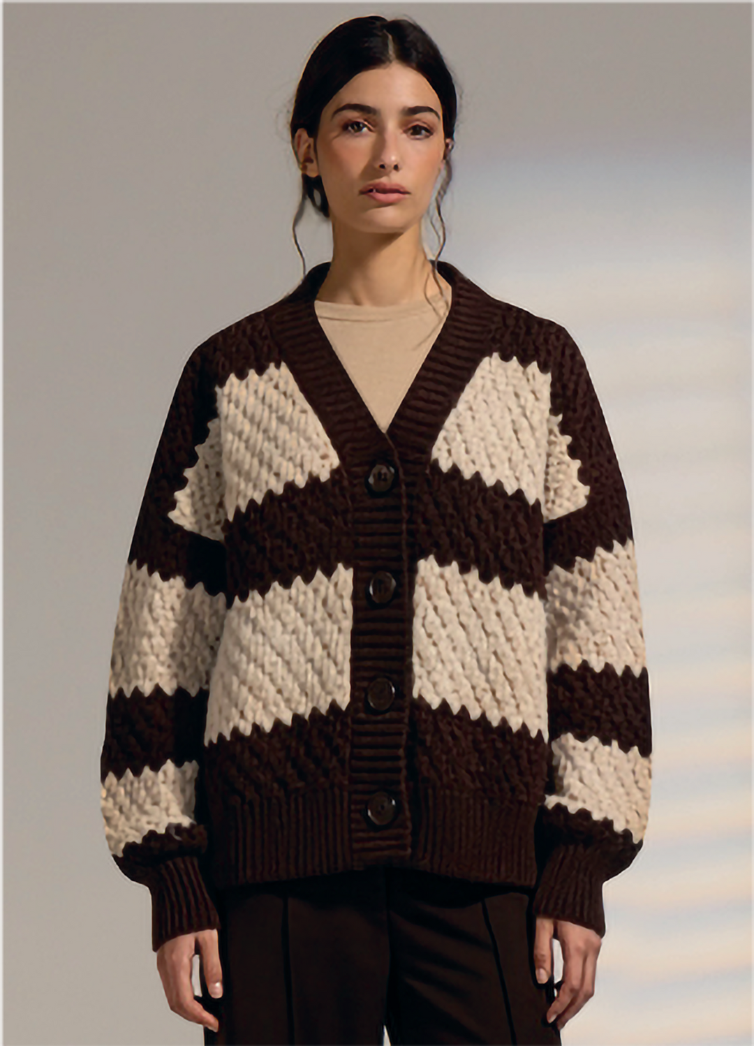 A woman modeling a chunky knit cardigan with bold brown and cream zigzag stripes, featuring a V-neck, button-front closure, and ribbed cuffs and hem.