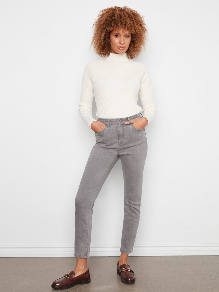 A woman modeling gray slim-fit jeans with a white ribbed mock neck top and brown leather loafers, standing against a white wall.