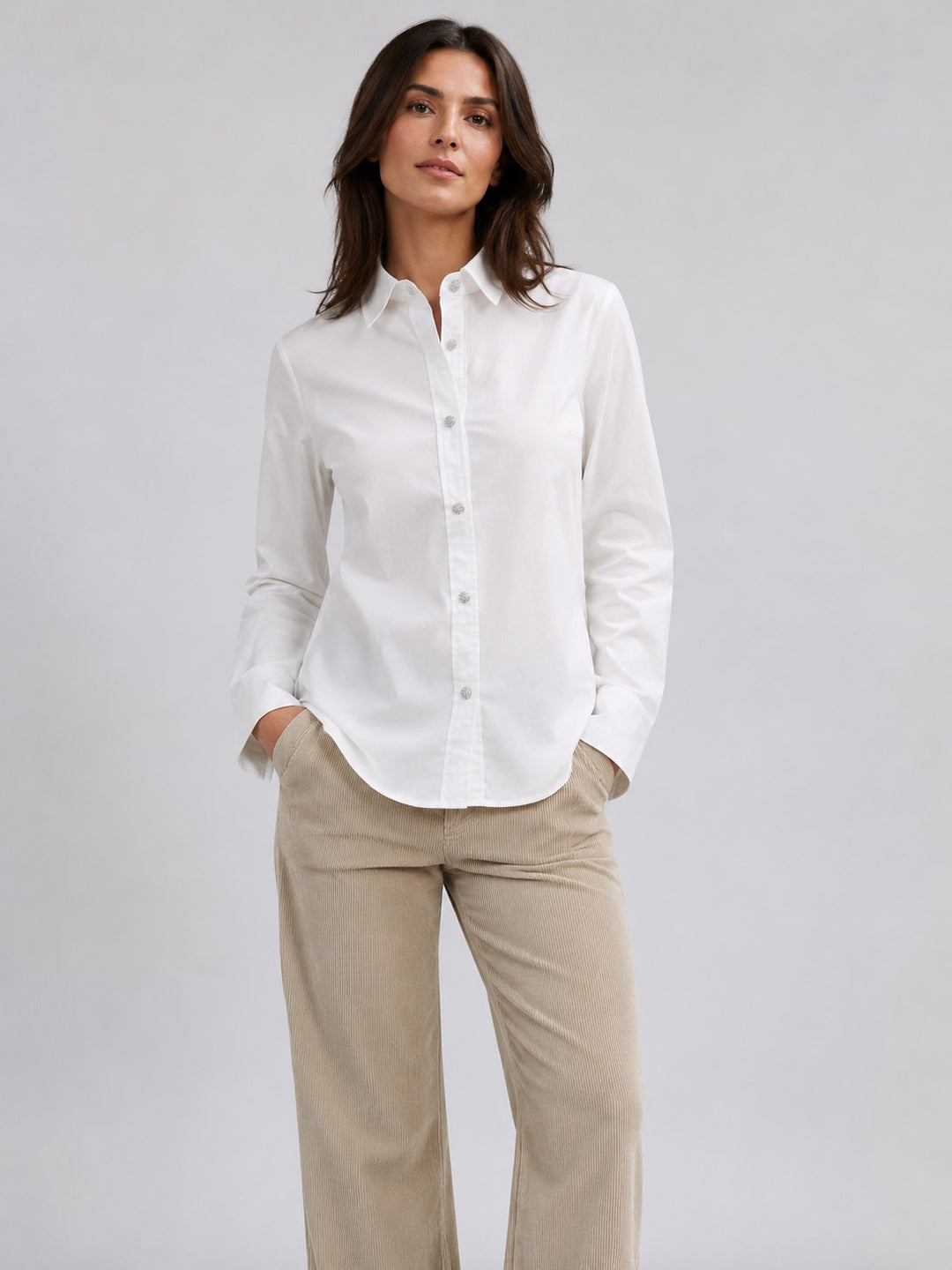 A woman modeling a white long-sleeve button-down shirt paired with beige trousers, standing against a neutral light gray studio background.