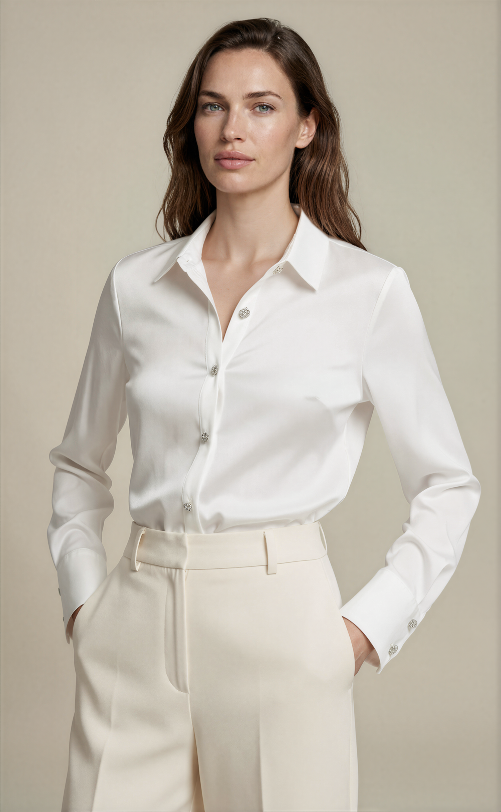 A woman modeling a white long-sleeve button-up shirt tucked into cream-colored trousers, set against a solid beige studio background.