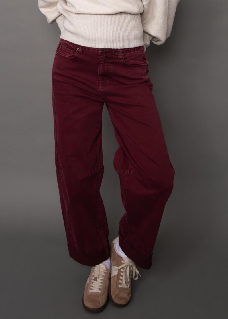 A person wearing loose-fitting burgundy corduroy pants with a cream ribbed sweater and beige sneakers, standing against a dark gray studio background.