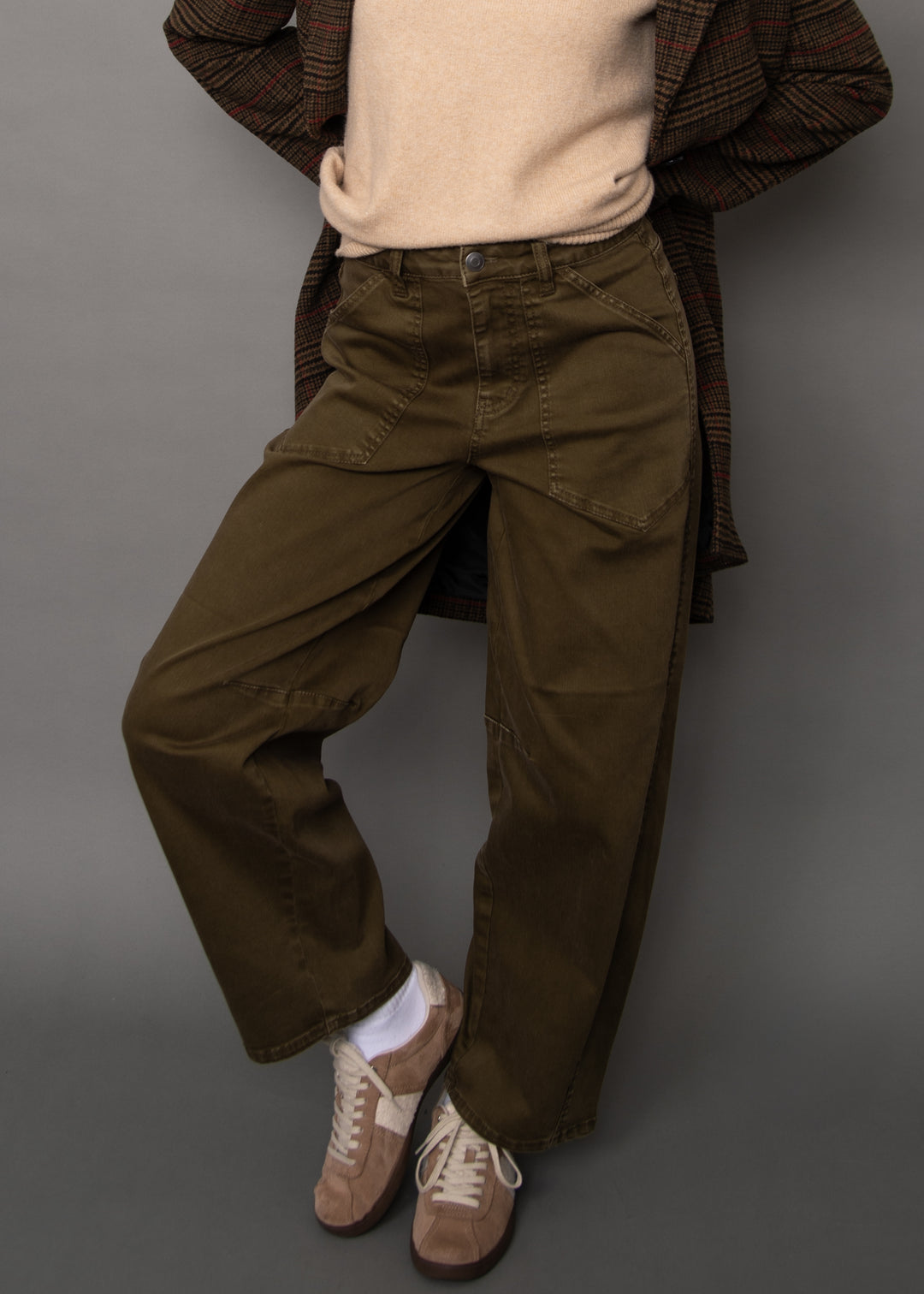 A person wearing olive green wide-leg cargo pants, styled with a beige sweater, a brown plaid coat, white socks, and brown sneakers against a solid gray studio background.