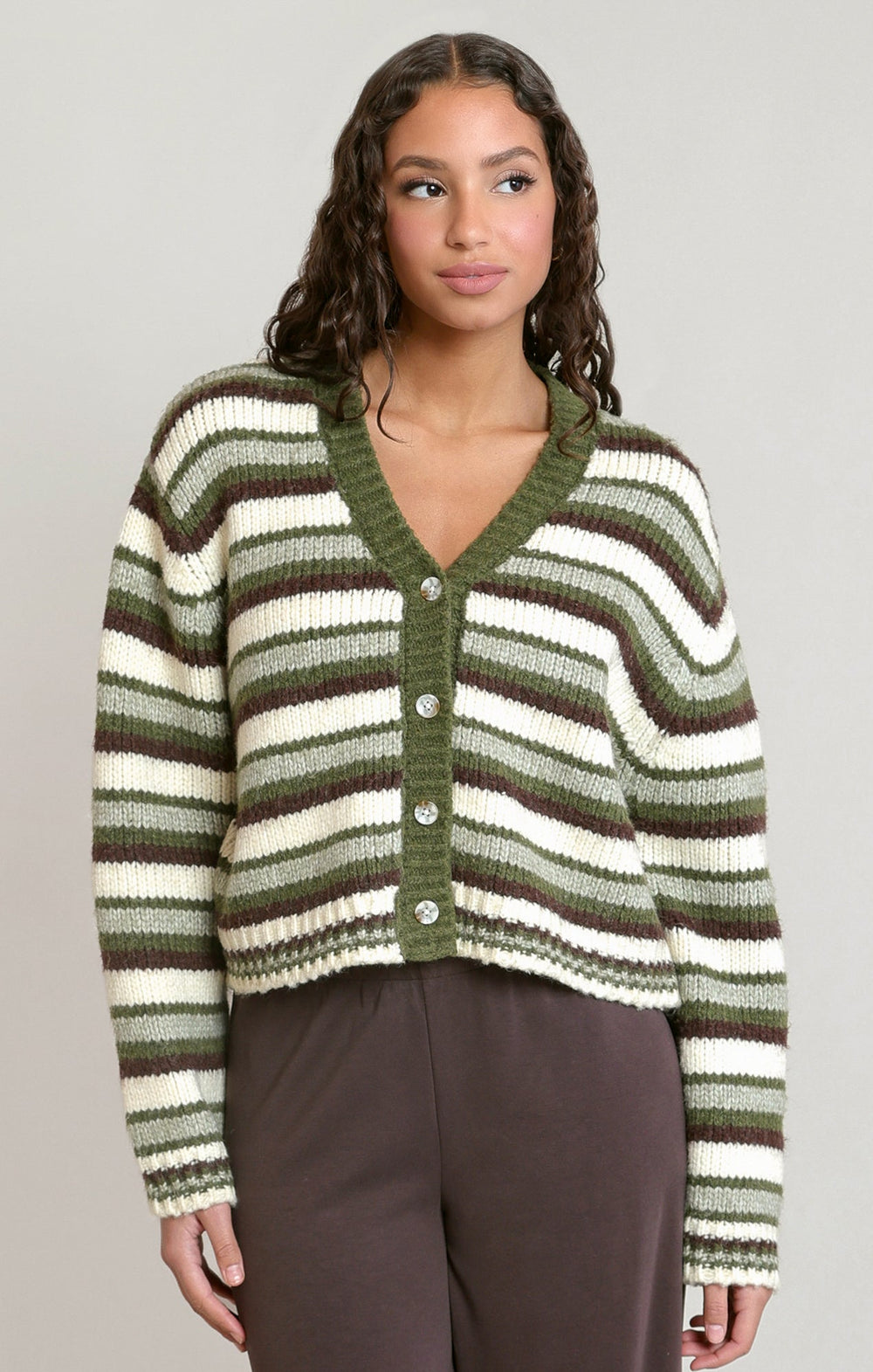 A woman wearing a cropped knit cardigan with horizontal olive green and cream stripes, paired with dark brown pants, against a solid light gray background.
