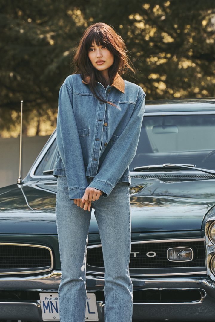 A woman wearing an oversized light blue denim jacket with a brown collar and matching light blue jeans, standing in front of a vintage dark green car outdoors.