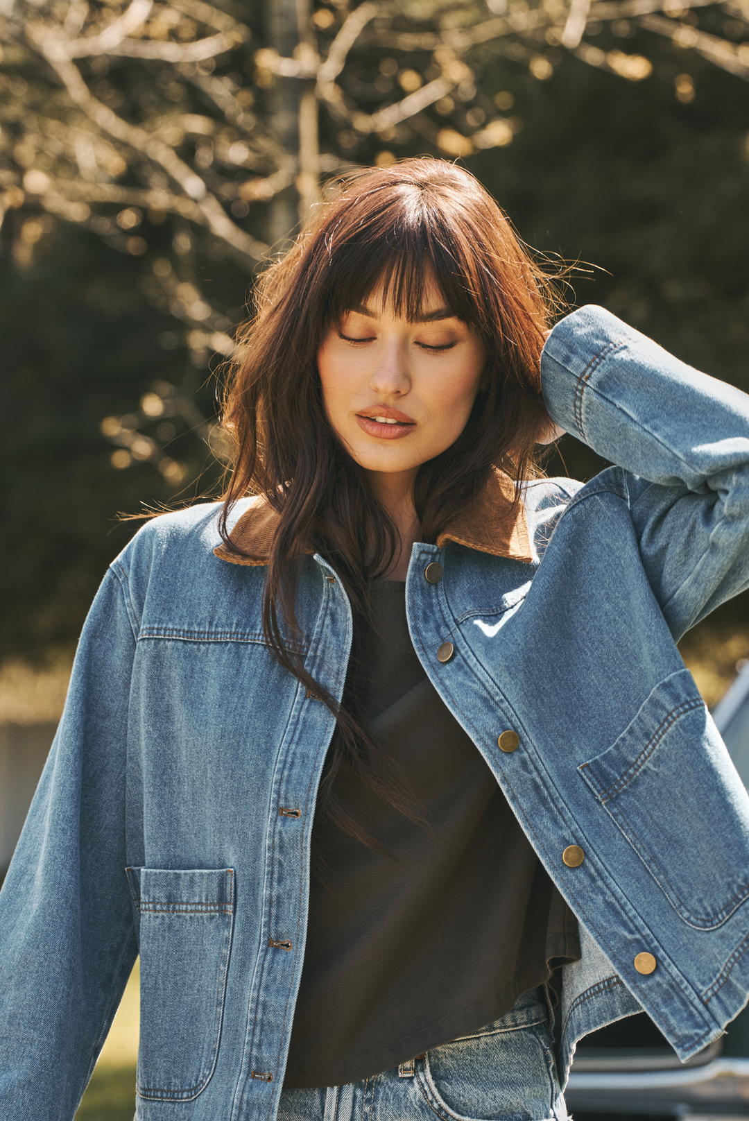 A woman wearing a light blue denim jacket with a brown corduroy collar over a black top, photographed outdoors with blurred trees in the background.